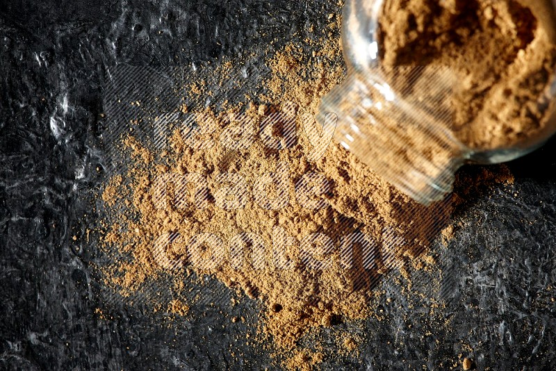 A flipped glass spice jar full of allspice powder and powder spilled out of it on a textured black flooring