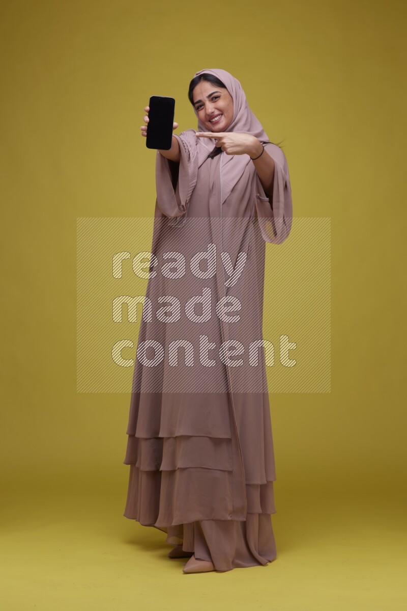 A woman Showing her Phone screen on a Yellow Background wearing Brown Abaya with Hijab