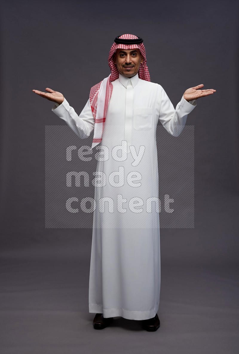 Saudi man wearing thob and shomag standing interacting with the camera on gray background