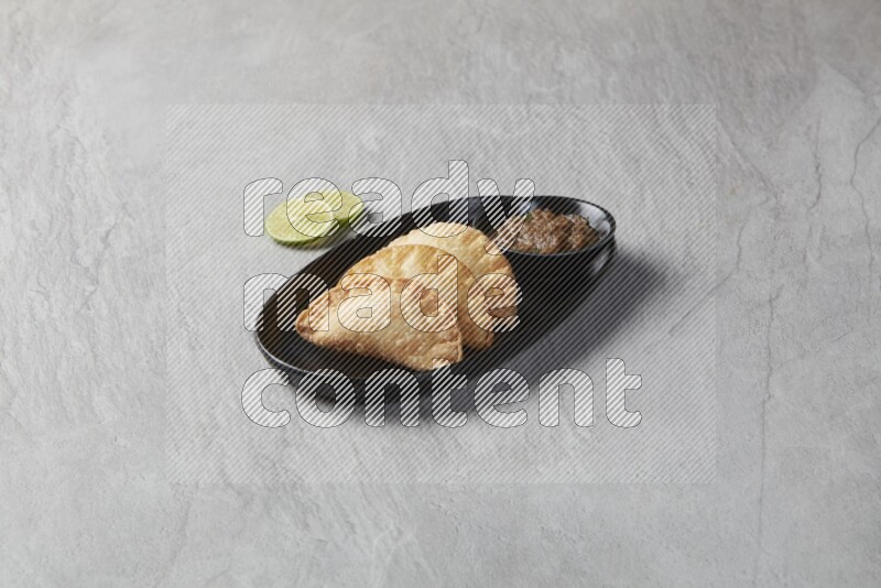 Three fried sambosas in an oval shaped black plate and a sauce in a black round ramekin with two lemon slices on the side on a gray background