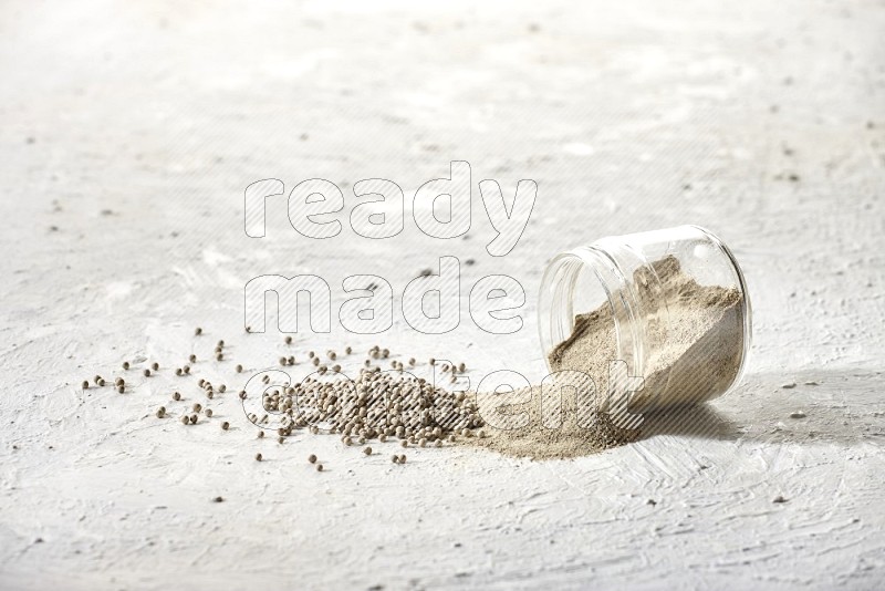 A flipped glass jar full of white pepper powder with spilled powder and white pepper beads on textured white flooring