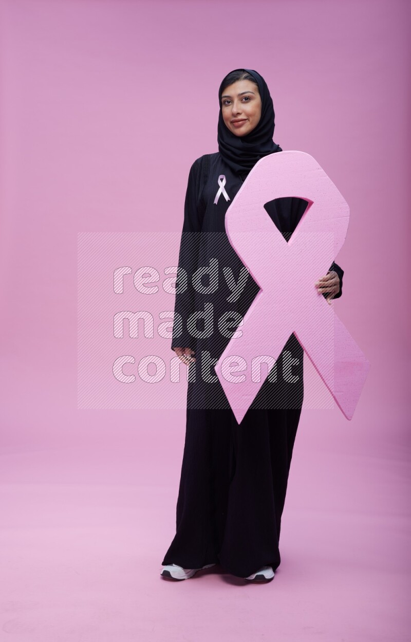 Saudi woman wearing pink ribbon on abaya standing holding awareness ribbon on plain pink background