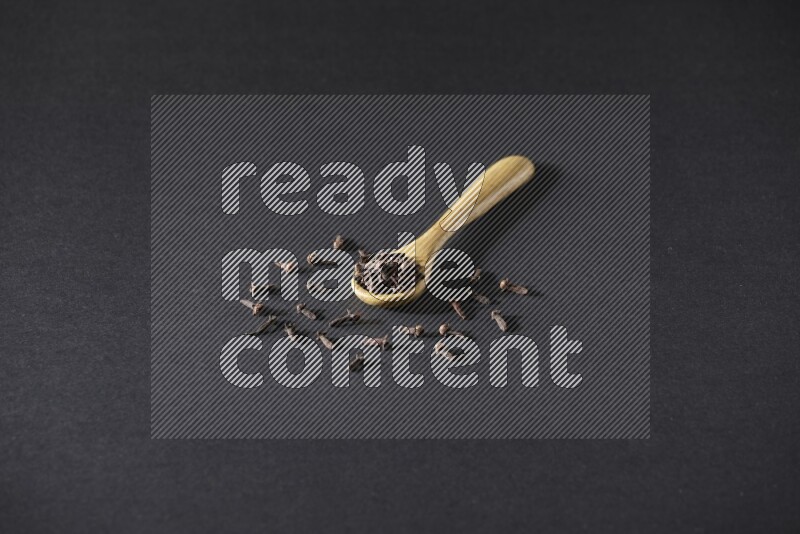 A wooden spoon full of cloves and some spreaded out of it on a black flooring