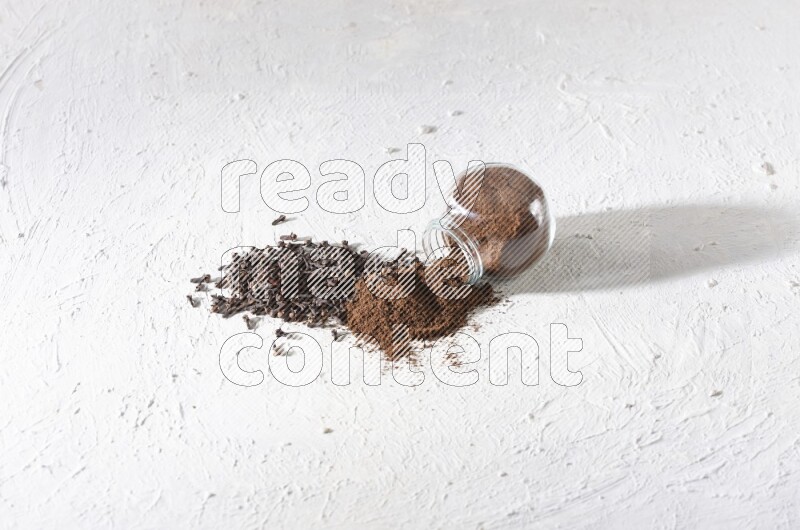 A glass spice jar full of cloves powder flipped and powder came out of it with cloves spread on textured white flooring