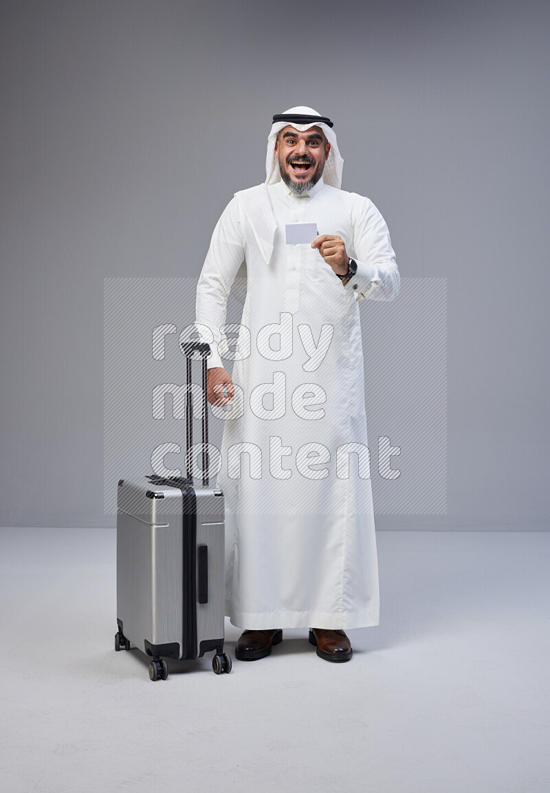 Saudi man wearing Thob and white Shomag standing holding Travel bag and ATM card on Gray background