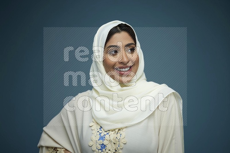 A Saudi woman posing in a blue background wearing an off-white Abaya with hijab