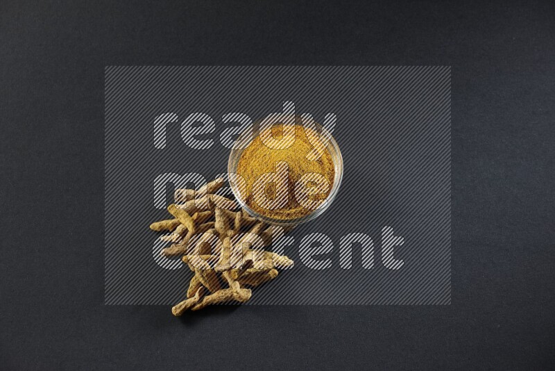 وعاء زجاجي ممتلئ ببودرة الكركم وأصابع الكركم المجففة بجانبه على خلفية سوداء