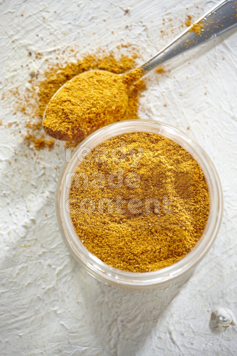 A glass jar and a metal spoon full of turmeric powder on a textured white flooring