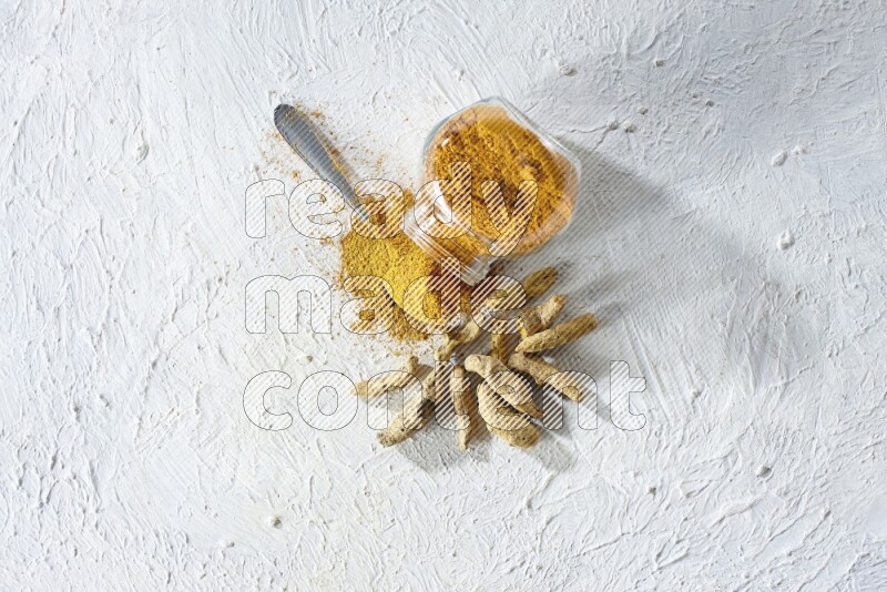 وعاء زجاجي للتوابل مقلوب وملعقة معدنية ممتلئة ببودرة الكركم والبودرة تناثرت منه مع أصابع الكركم المجففة على خلفية بيضاء