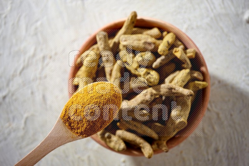A wooden spoon full of turmeric powder above a wooden bowl full of dried turmeric whole fingers on a textured white flooring