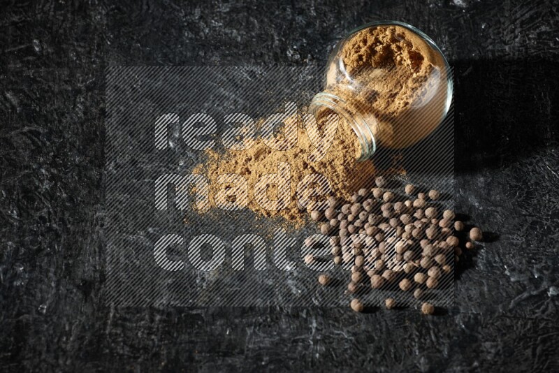 وعاء زجاجي للتوابل مقلوب ممتلئ ببودرة بهار الفلفل الحلو والبودرة تناثرت منه مع الحبوب على خلفية سوداء