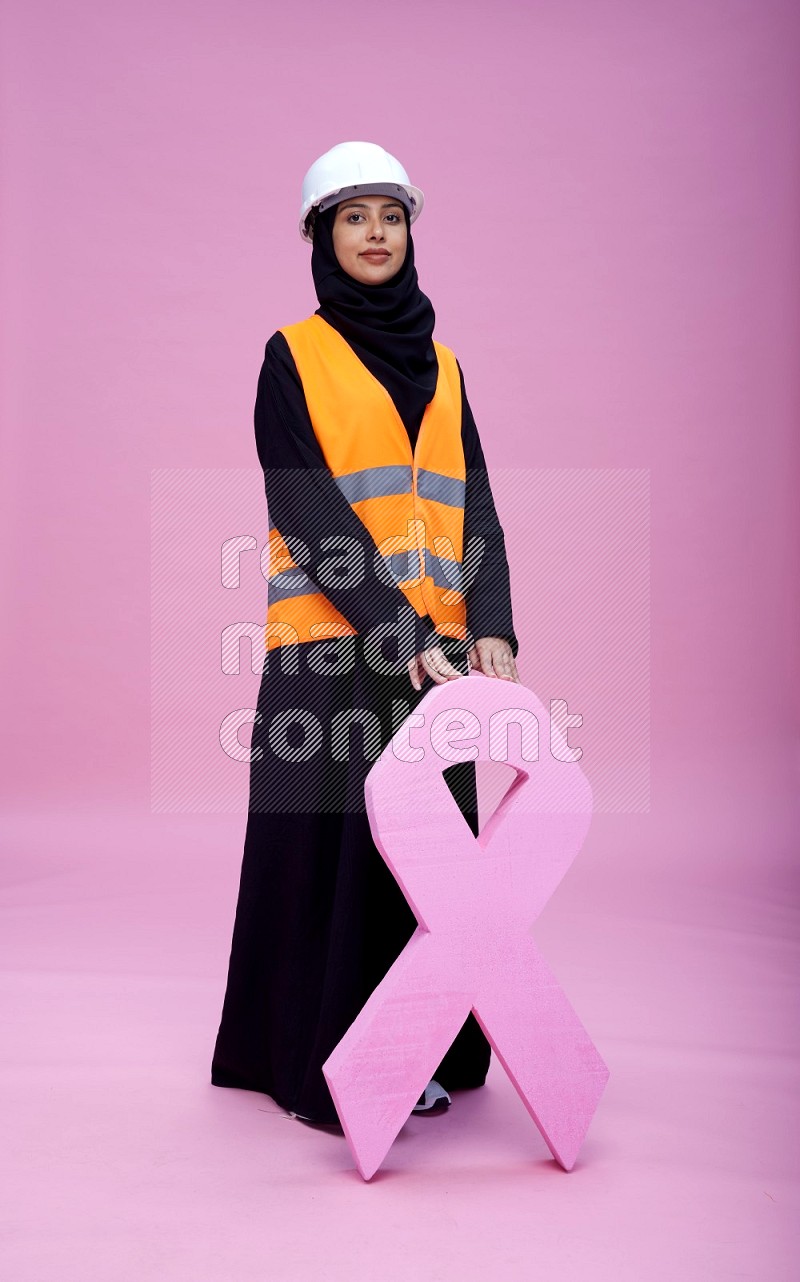 Saudi woman wearing Abaya with engineer vest and helmet standing awareness ribbon on pink background