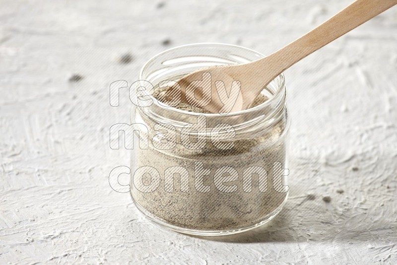 A glass jar and wooden spoon full of white pepper powder on textured white flooring