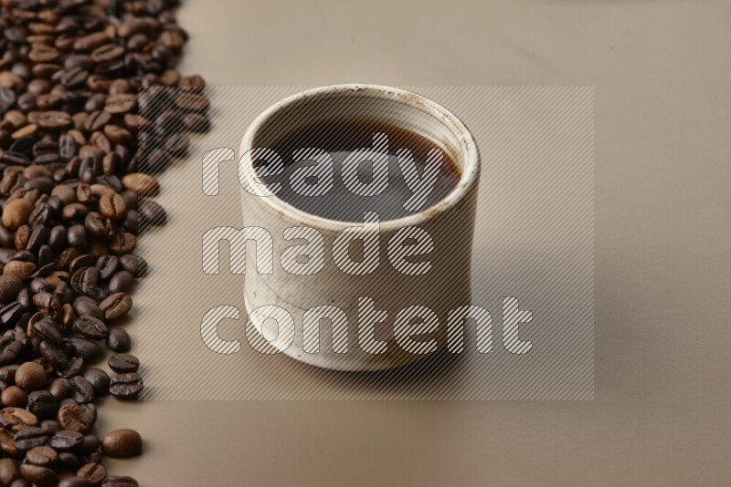 A beige pottery cup of coffee surrounded by roasted coffee beans on beige background