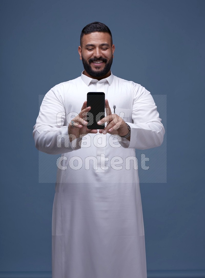 A man Showing his phone in Blue Background wearing Saudi Thob