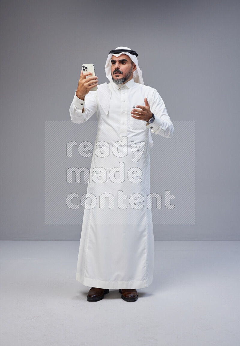 Saudi man Wearing Thob and white Shomag standing texting on phone on Gray background