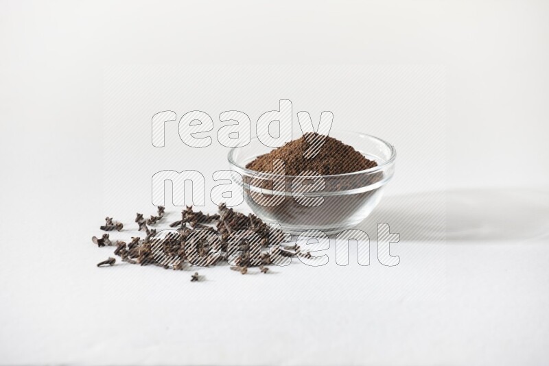 A glass bowl full of cloves powder and cloves grains spread on white flooring