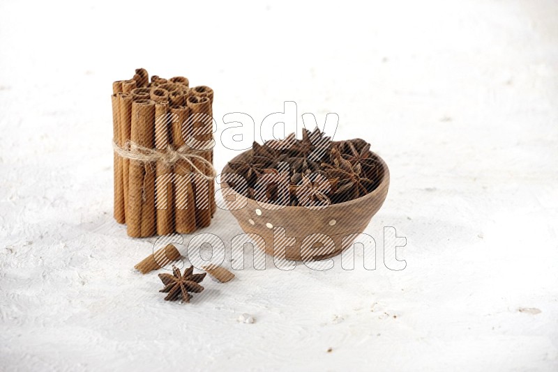A stacked and bounded cinnamon sticks and a wooden bowl full of star anise on a white background