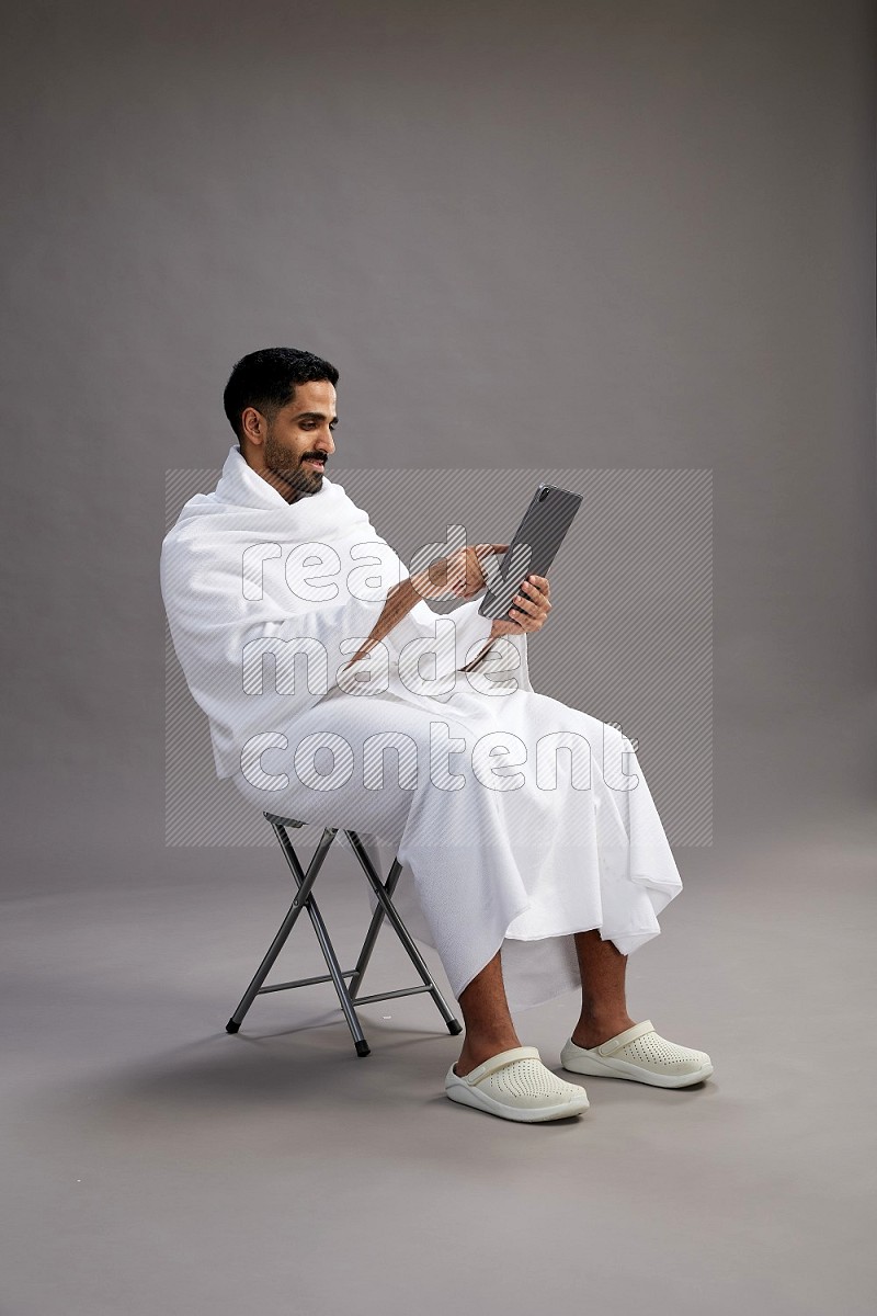 A man wearing Ehram sitting on chair holding a tablet on gray background