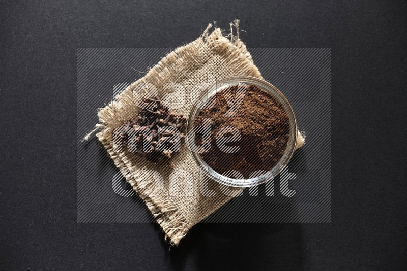 A glass bowl full of cloves powder with cloves grains on a burlap piece on a black flooring