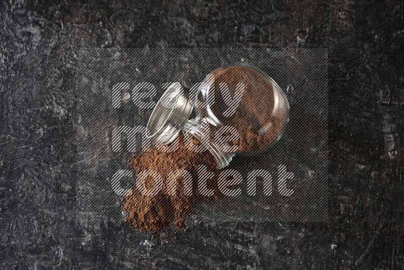 A flipped glass spice jar full of cloves powder and powder came out of it on textured black flooring