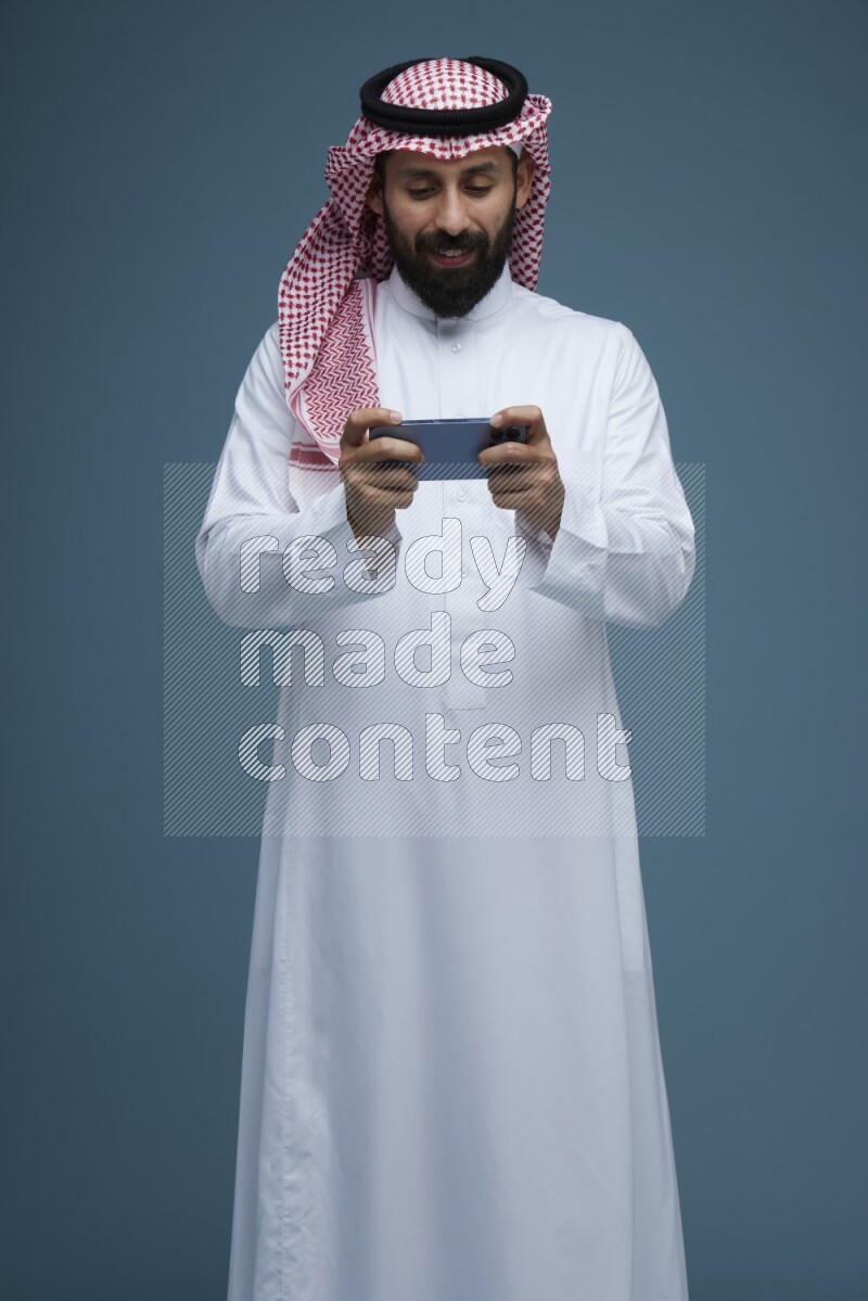 A man Playing a Game on his phone in a blue background wearing Saudi Thob with Shomag
