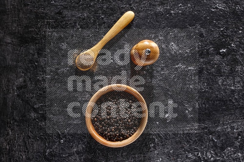 A wooden bowl full of black pepper and a wooden spoon full of black pepper powder and a wooden grinder on a textured black flooring