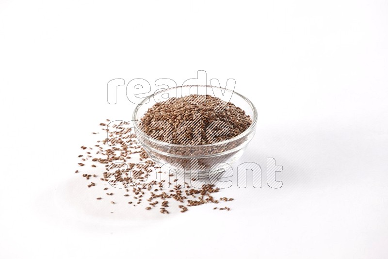 A glass bowl full of flax seeds surrounded by flax seeds on a white flooring