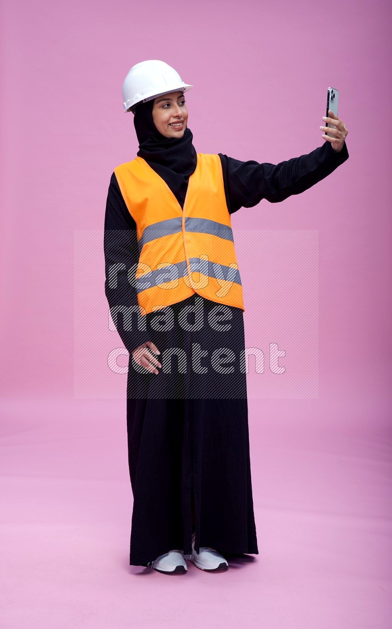 Saudi woman wearing Abaya with engineer vest and helmet standing taking selfie on pink background