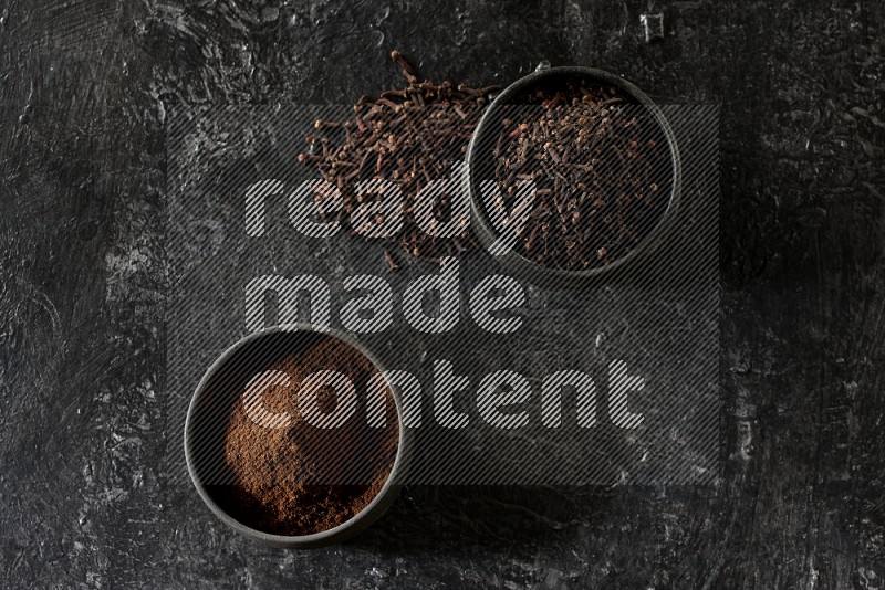 2 Black pottery bowls full of cloves and the other full of cloves powder on textured black flooring