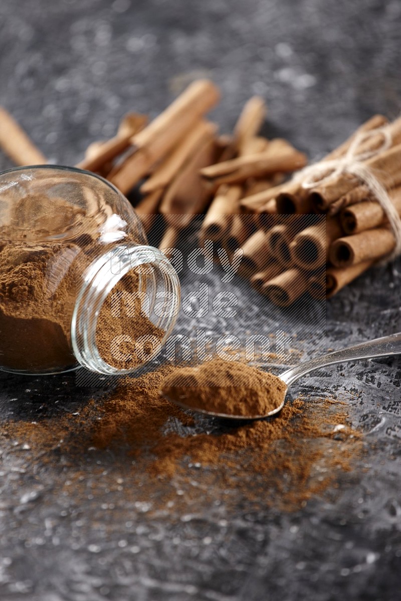 Herbal glass jar full cinnamon powder flipped and a metal spoon full of powder, cinnamon sticks stacked and bounded in the back on textured black background in different angles