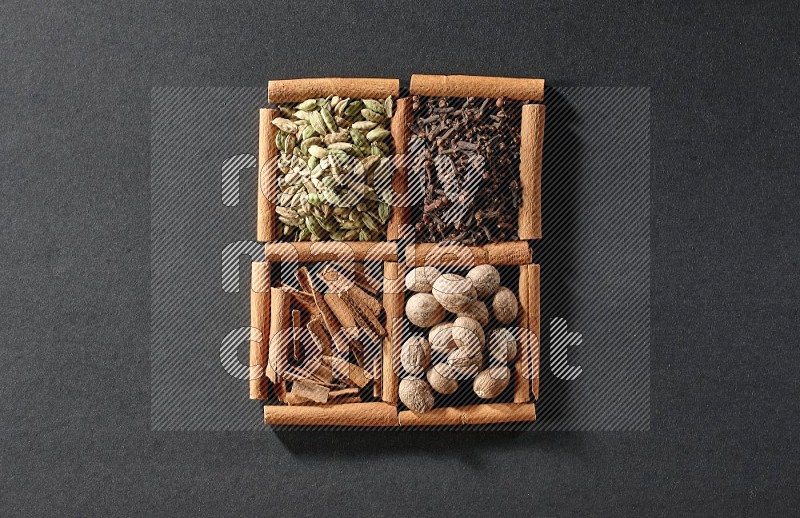 4 مربعات من عيدان القرفة ممتلئة بقطع القرفة وحبوب الهيل والقرنفل وجوزة الطيب على أرضية سوداء