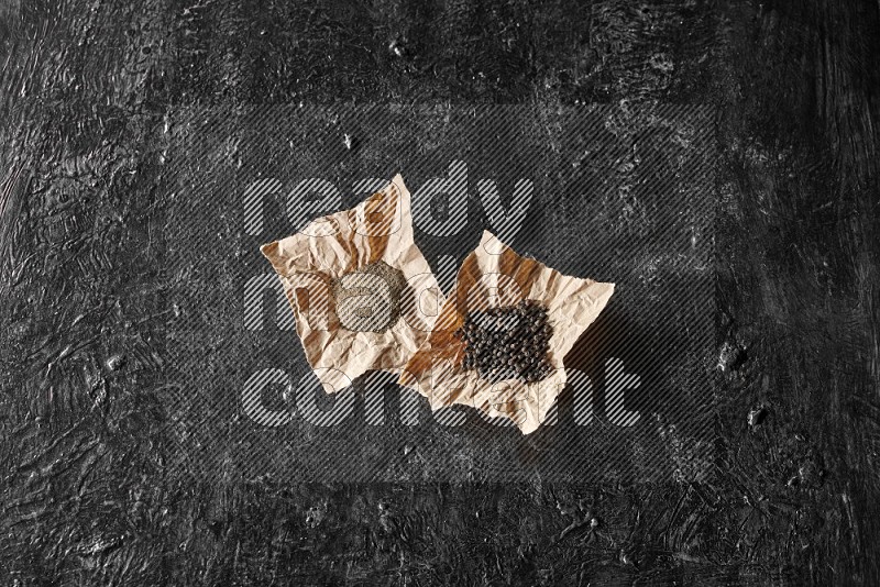 Black pepper and black pepper powder on 2 crumpled paper on a textured black flooring