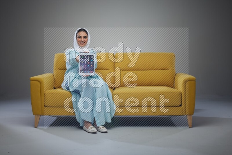 A Saudi woman wearing a light blue Abaya and white head scarf sitting on a yellow sofa and showing her tablet's screen while pointing to it eye level on a grey background