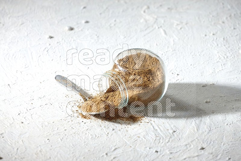 A flipped glass spice jar and a metal spoon full of cumin powder and powder spilled out on textured white flooring