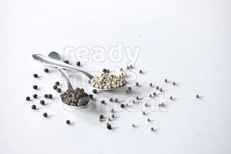 2 metal spoons full of black and white pepper beads with spreaded beads on white flooring