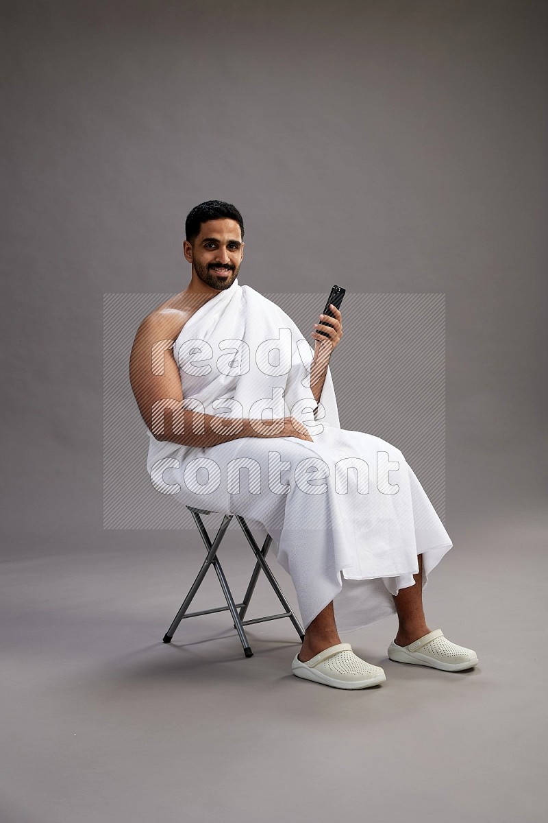 A man wearing Ehram sitting on chair texting on phone on gray background