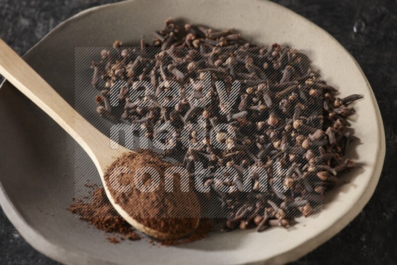 A Pottery plate full of cloves and a wooden spoon full of cloves powder on it on a textured black background