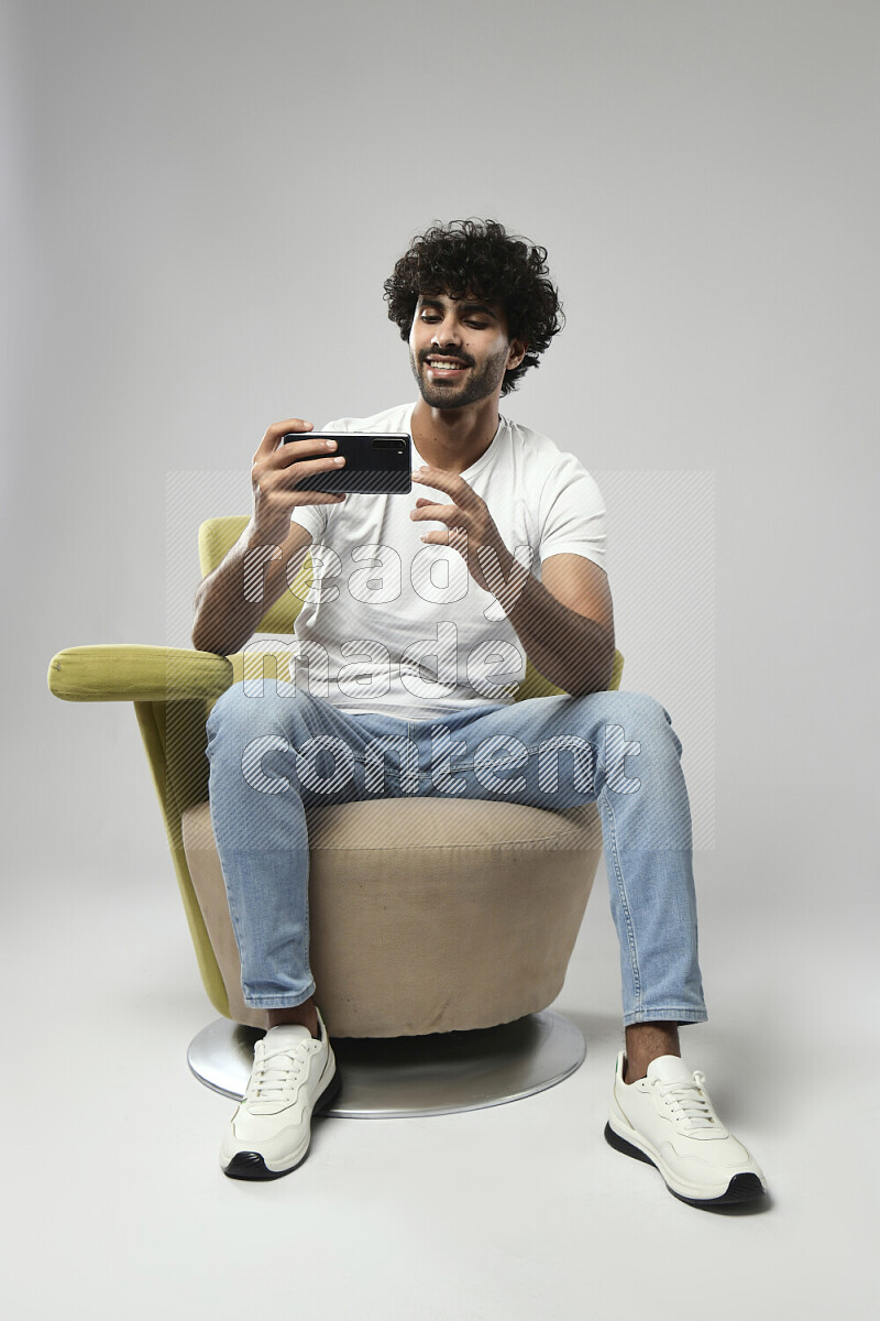 رجل يرتدي ملابس كاجوال يجلس علي كرسي و يصور بهاتفه علي خلفية بيضاء