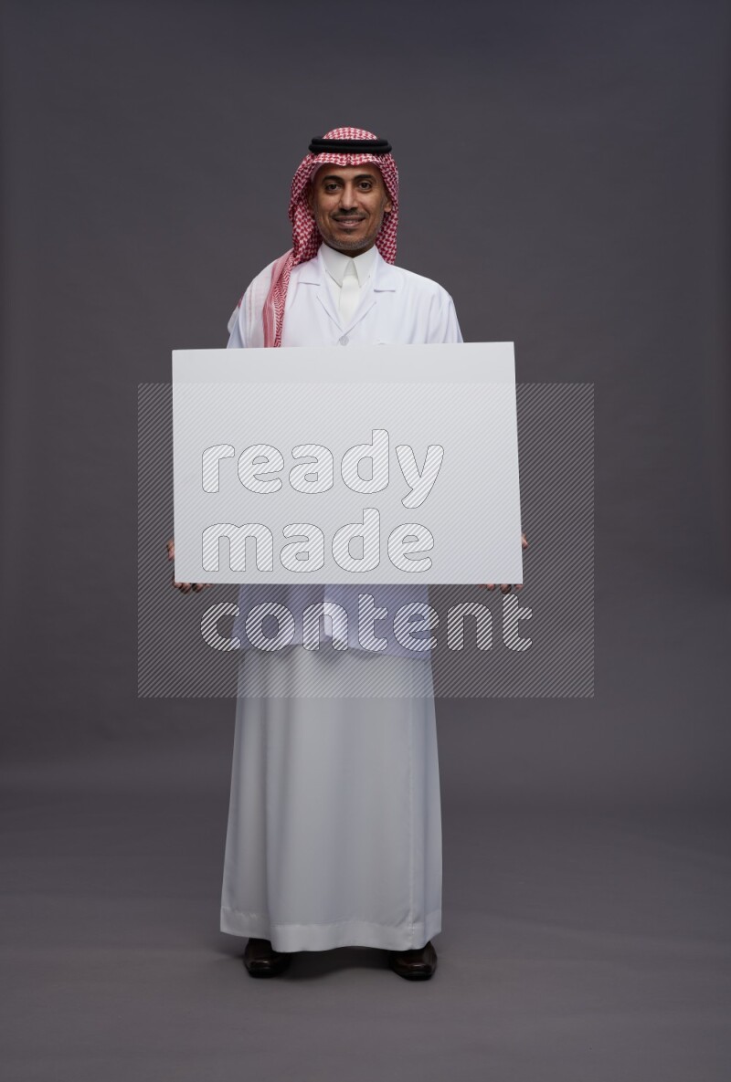 Saudi man wearing thob with lab coat and shomag with pocket employee badge standing holding board on gray background