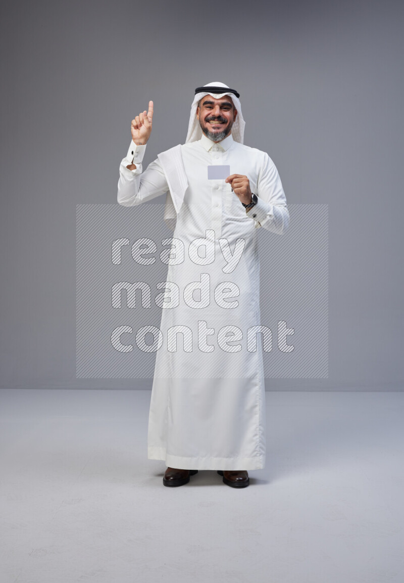 Saudi man Wearing Thob and white Shomag standing holding ATM card on Gray background