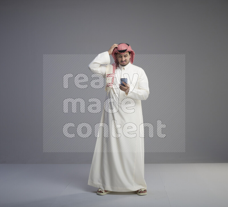 A  Saudi man standing wearing thob and red shomag texting on phone on gray background