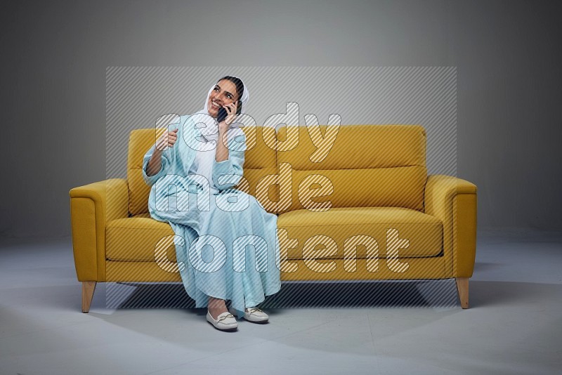 A Saudi woman wearing a light blue Abaya and a white head scarf sitting on a yellow sofa and talking in the phone while making different poses eye level on a grey background