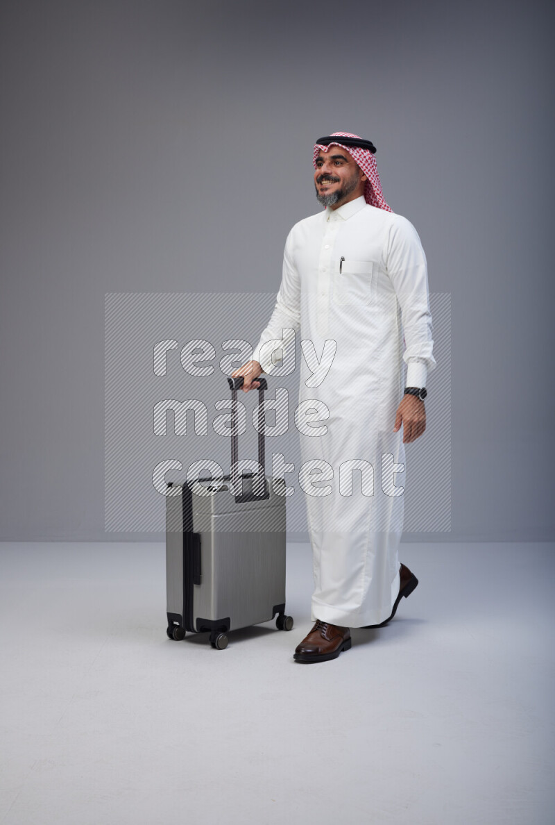 Saudi man wearing Thob and red Shomag standing holding Travel bag on Gray background