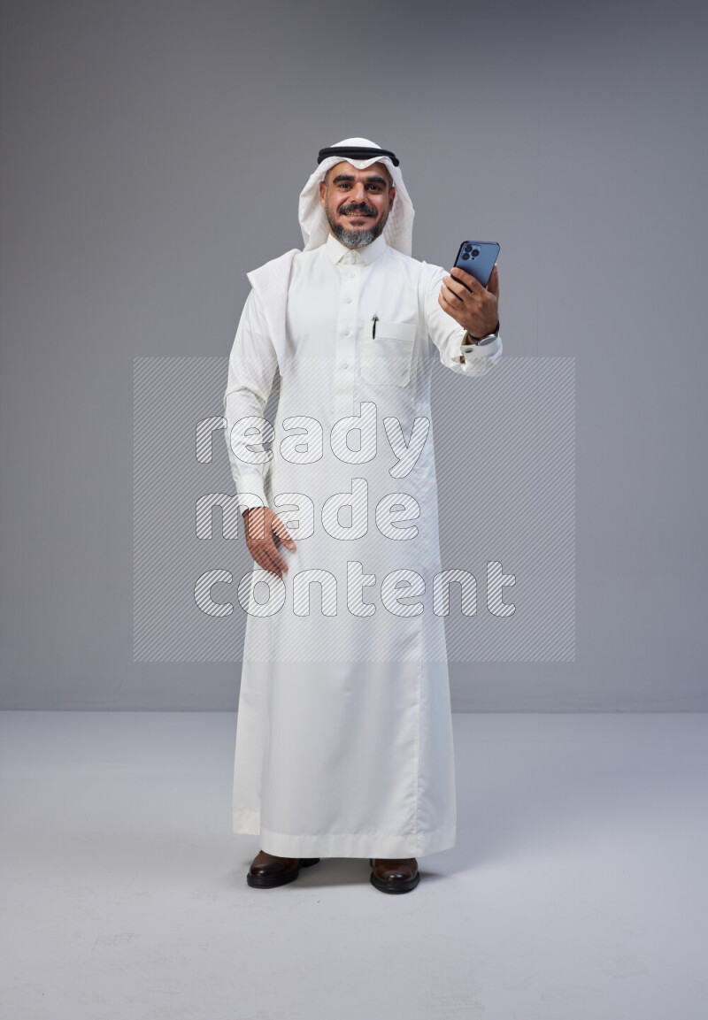 Saudi man Wearing Thob and white Shomag standing taking selfie on Gray background