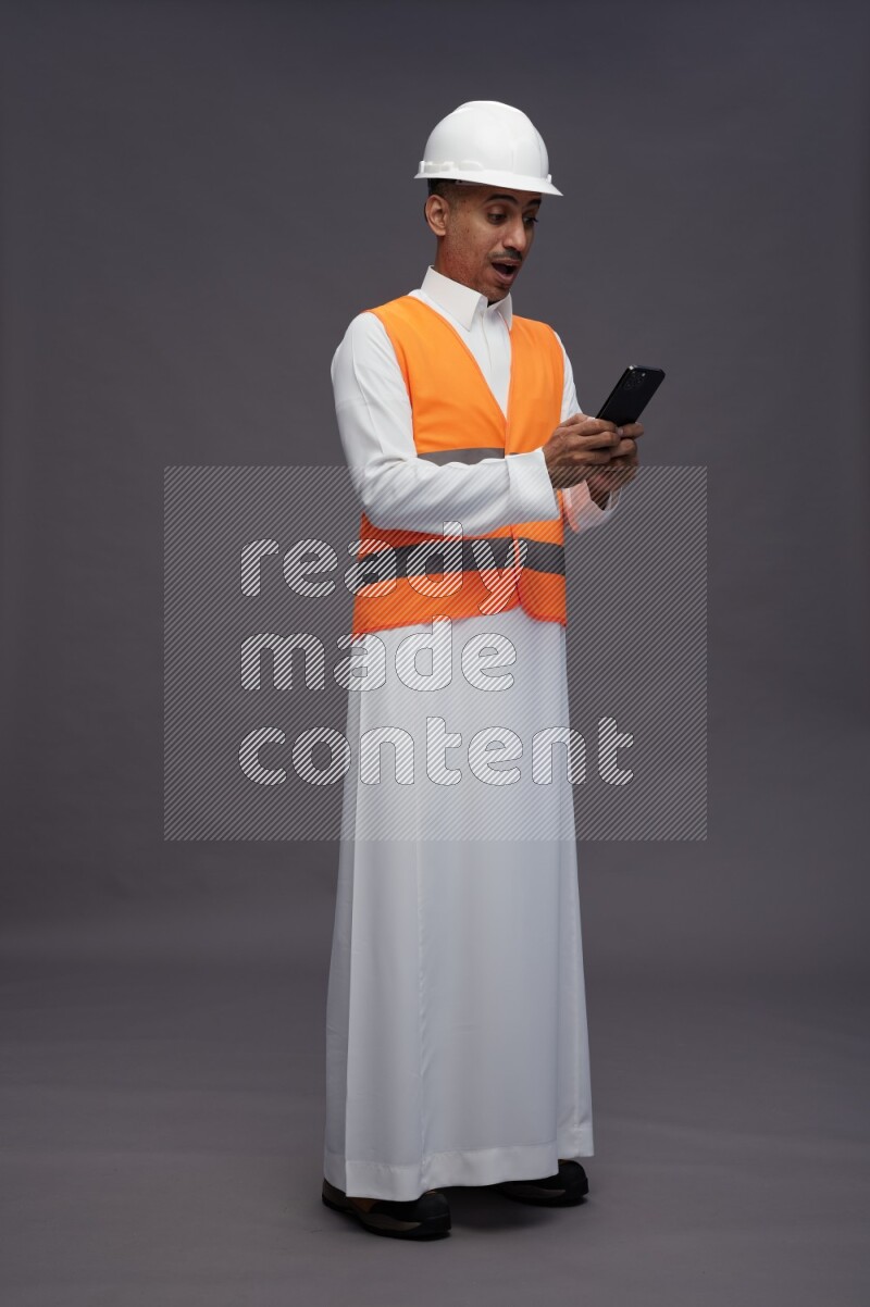 Saudi man wearing thob with engineer vest standing texting on phone on gray background