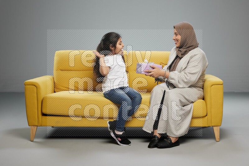 A girl sitting on a yellow sofa and giving a gift to her mother on gray background