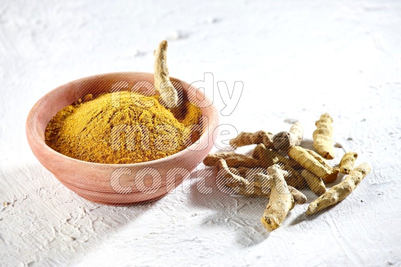 A wooden bowl full of turmeric powder and dried turmeric whole fingers beside it on textured white flooring