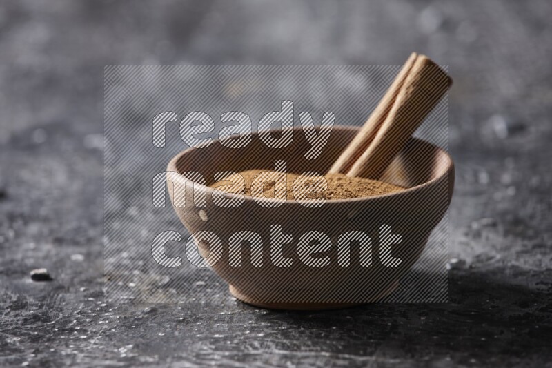 Wooden bowl full of cinnamon powder and a cinnamon stick on a textured black background