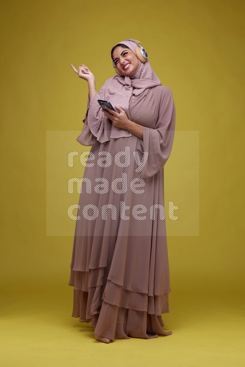 A woman Listening to Music on a Yellow Background wearing Brown Abaya with Hijab